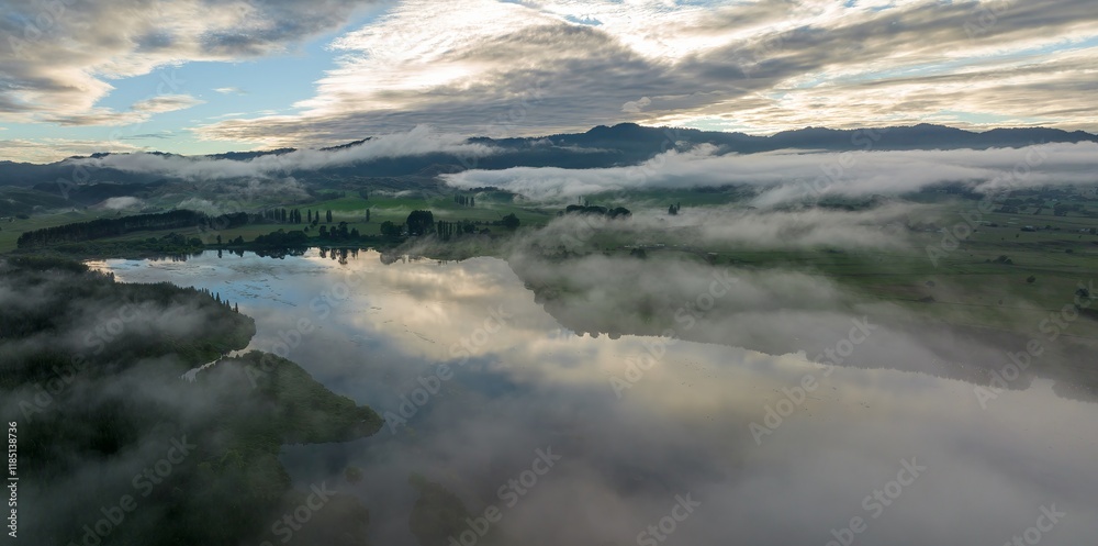 Misty morning over a tranquil lake. Reflections of clouds on still water. Peaceful rural landscape. LAKE ANIWHENUA, MURUPARA, BAY OF PLENTY, NEW ZEALAND Misty morning over a tranquil lake. Reflections of clouds on still water. Peaceful rural landscape. LAKE ANIWHENUA, MURUPARA, BAY OF PLENTY, NEW ZEALAND