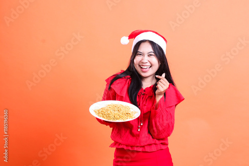 portrait of a woman wearing Christmas clothes, Santa hat, cheerful to the camera, hands holding a white plate filled with noodles to the right and a love sign saranghaeyo, isolated background photo