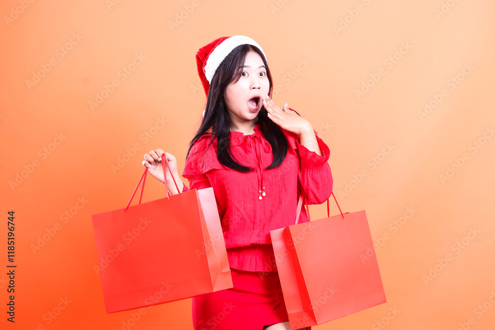 Fototapeta premium teenage woman charm wearing red dress and santa hat, standing sideways left surprised to left hand lifting red bag of christmas gifts and covering mouth, isolated on orange background