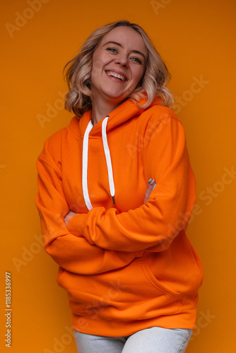 A smiling woman in an orange hoodie with crossed arms, standing in front of a vibrant yellow background. She appears confident and cheerful, representing positive energy and casual fashion. photo