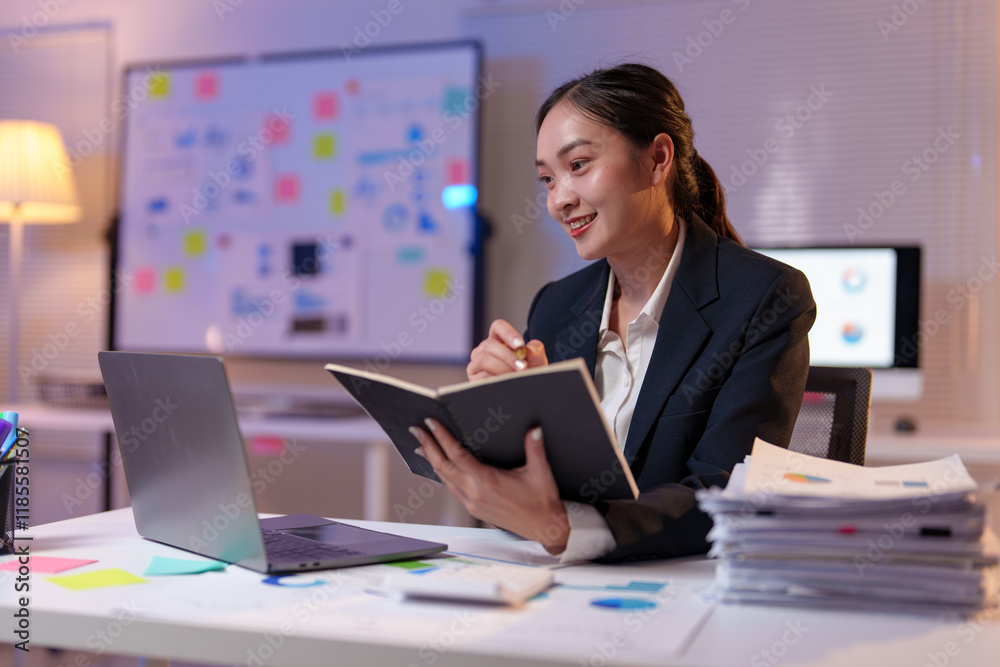 Young professional woman writing on a notebook and smiling while working late at night in a modern office with a laptop and documents on the desk Young professional woman writing on a notebook and smiling while working late at night in a modern office with a laptop and documents on the desk