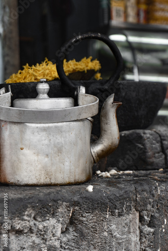 Aluminum tea kettle sitting on the grey stone platform in Indian traditional chai stall. Indian tea shop with big black cauldron with fried snacks in the back. Roadside shop traditional Indian snacks