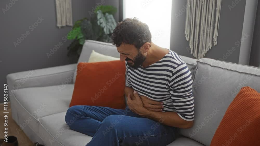 Young man with beard sitting on couch in living room clutching stomach in pain Young man with beard sitting on couch in living room clutching stomach in pain