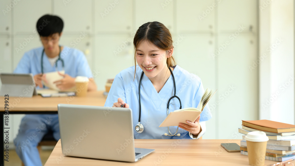Smiling medical student in scrubs holding a book and studying on her laptop Smiling medical student in scrubs holding a book and studying on her laptop
