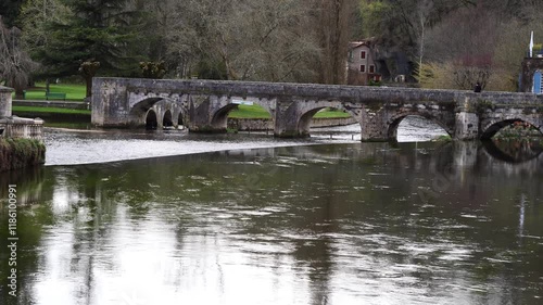 A bridge spans a river with a green bridge in the background. The bridge is old and has a stone structure.
