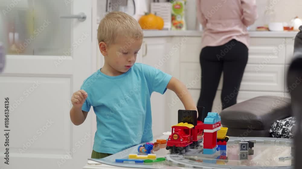 Young boy stands at glass table focused on building train track out of blocks. Boy hand is reaching for next piece of track. In background adult female stands near cabinet. Focus, joy, and creativity.