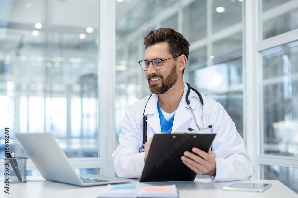 Smiling doctor records patient data using laptop for video call, man in white medical coat working inside modern clinic office at workplace, consulting patients remotely.