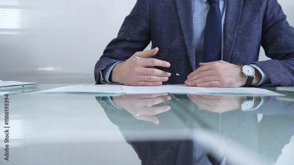 Businessman examining contract terms, using a pen to gesture, in a contemporary office environment, with reflection on a glass desk. Business people or audit concept