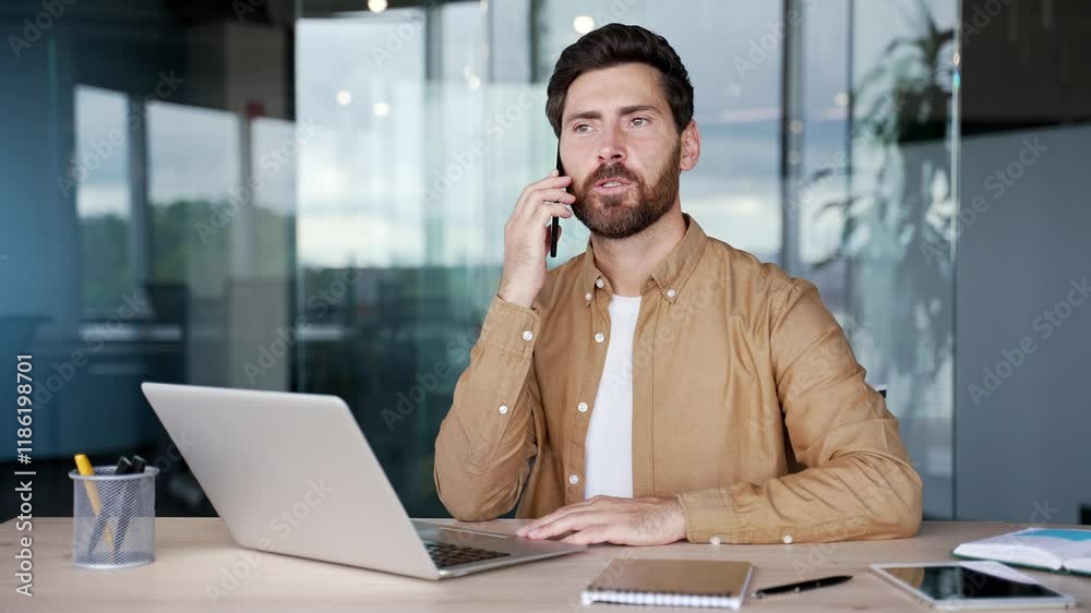 Confident businessman talking on mobile phone while sitting at workplace in modern office. Handsome bearded manager communicates with a client or conducts a conversation with a business partner Confident businessman talking on mobile phone while sitting at workplace in modern office. Handsome bearded manager communicates with a client or conducts a conversation with a business partner