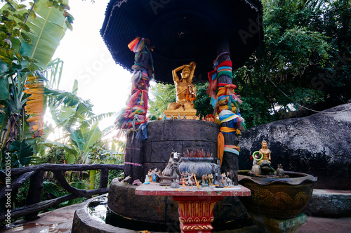 A golden Buddha statue under a canopy, surrounded by colorful cloths and smaller statues, set in a lush garden with tropical plants. photo