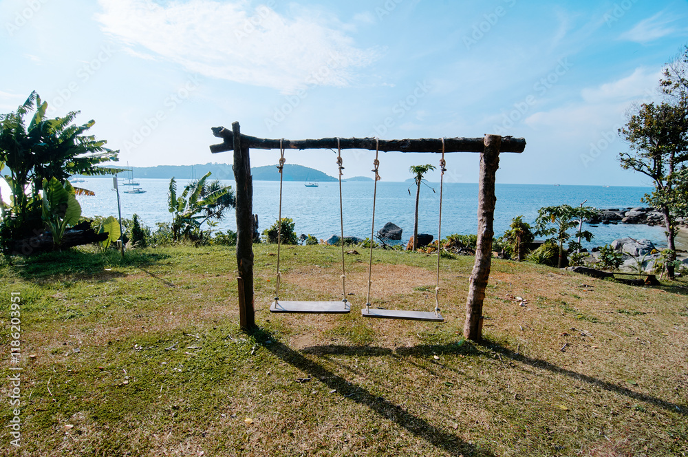 A rustic wooden swing set with two swings overlooking a scenic ocean view, surrounded by lush greenery and clear blue sky. A rustic wooden swing set with two swings overlooking a scenic ocean view, surrounded by lush greenery and clear blue sky.
