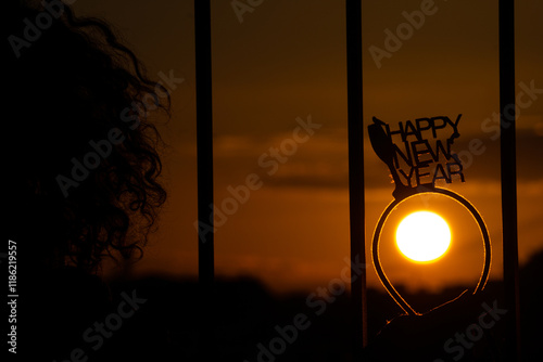 Sagoma di una donna riccia al tramonto, che tiene in mano un cerchietto con scritto happy new year, che contorna il disco del sole arancione photo