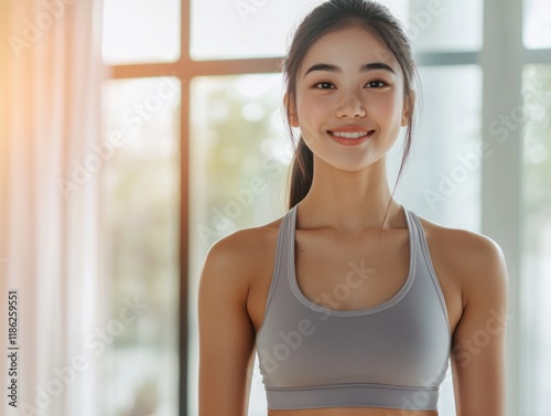 Free Download Young Asian woman in gray sports bra smiling during indoor exercise session with bright natural light and large empty space for text. - GFXLoad