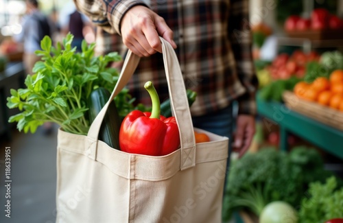 Person holds reusable produce bag filled with fresh veggies at local market. Bag beige canvas. Red bell peppers zucchini, leafy greens inside. Person wearing plaid shirt. Blurred background shows photo