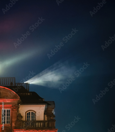 Illuminated stage lights and projected beams create an ethereal glow against a historical building, blending architectural beauty with artistic lighting effects.