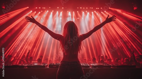 A DJ stands confidently behind the turntables, arms raised high while engaging with a cheering crowd. Red lights fill the venue, creating a dynamic atmosphere. photo