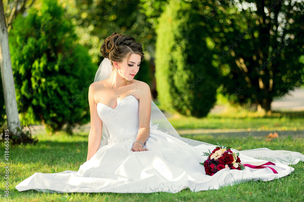 A woman in a white wedding dress sits on the grass with a bouquet of red flowers in her lap A woman in a white wedding dress sits on the grass with a bouquet of red flowers in her lap
