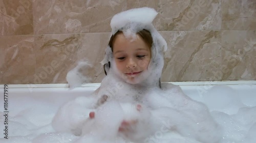 A little girl washes and bathes in the bathroom with foam smiling. Funny baby face bathing in a bathtub. Foam on the head.