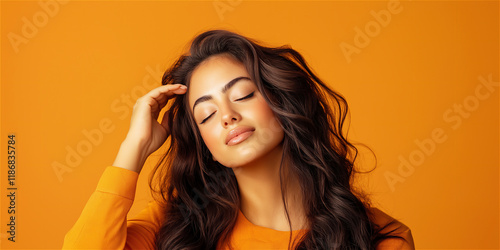 Confident woman with dark, voluminous wavy hair standing against a bright orange background, gently touching her face and smiling, embracing positivity. photo