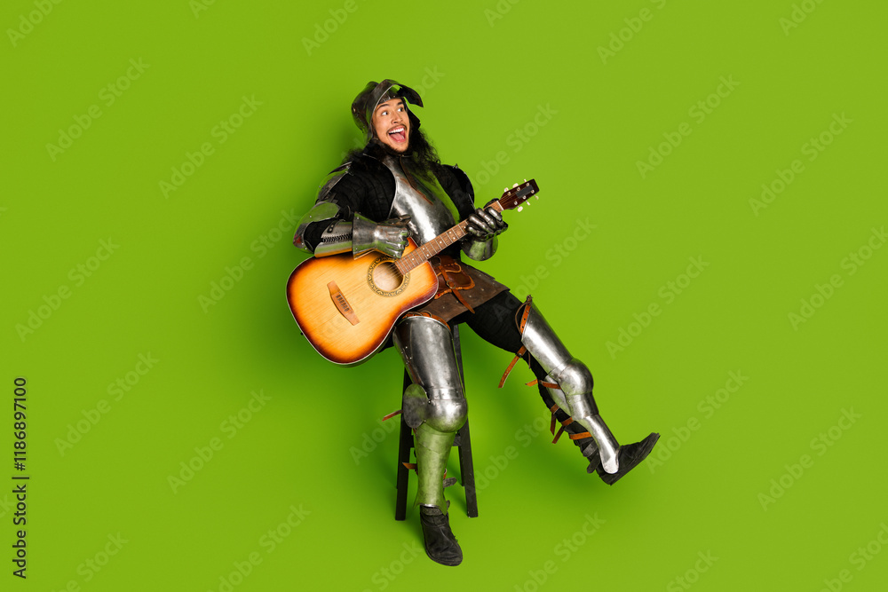 A young knight dressed in medieval armor joyfully playing an acoustic guitar against a vibrant green background A young knight dressed in medieval armor joyfully playing an acoustic guitar against a vibrant green background