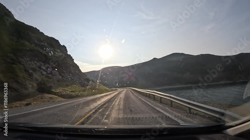 Driving through tundra on the E69 road in northern Norway. Rocky hills with grass surround the route. A sea bay appears on the right. The sun shines ahead as the car enters the Nordkapp tunnel. photo