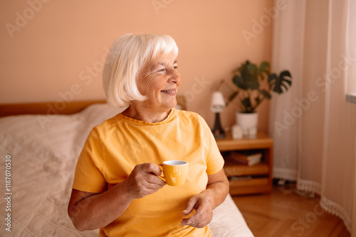 Happy Caucasian blonde senior woman sitting on a white bed with a cup in her hand. Female relaxing and drinking cup of hot coffee or tea in the bedroom.