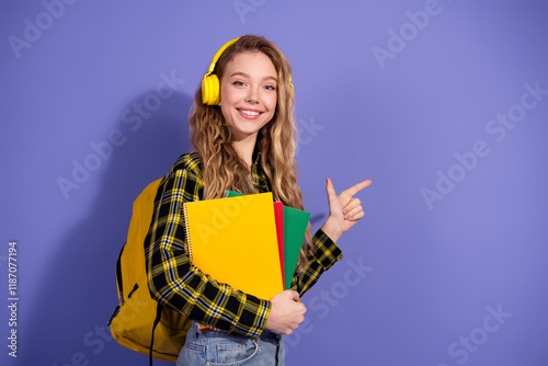Cheerful young female student indicating direction, wearing headphones and carrying colorful notebooks on a blue background