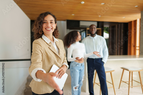 Female real estate professional in beige blazer presents property details to diverse couple during consultation in contemporary residential space with wooden accents.