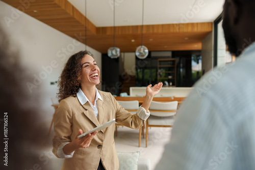 Caucasian female real estate professional in beige blazer enthusiastically discusses property details with diverse clients using digital tablet in modern office setting.