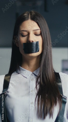 Vertical Screen: Young professional woman standing in an office corridor with her mouth taped shut by a man's hands, symbolizing censorship or workplace inequality. Concept of oppression and silencing