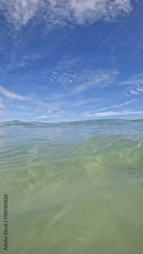 Transparent waves roll in, the camera goes under water and opens a beautiful view of the clean sandy bottom. In the background is a blue sky. Video vertical 4k