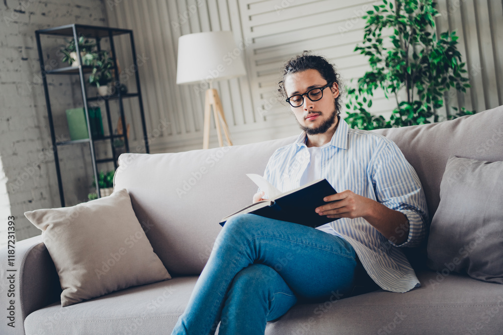 Obraz premium Handsome young man relaxing at home on a sofa while reading a book in a comfortable and well-lit interior