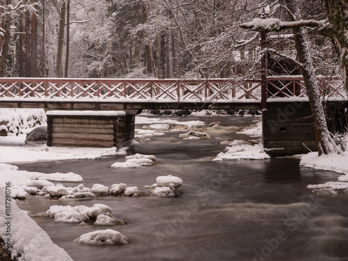 A secluded winter landscape with a rushing stream or waterfall amidst snowy banks. A vintage bridge is visible. The atmosphere of harmony and silence of the forest’s winter nature, cozy tranquility, l photo