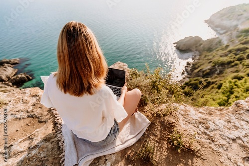 A woman is sitting on a rock overlooking the ocean with a laptop in front of her. She is enjoying the view and the peacefulness of the location.