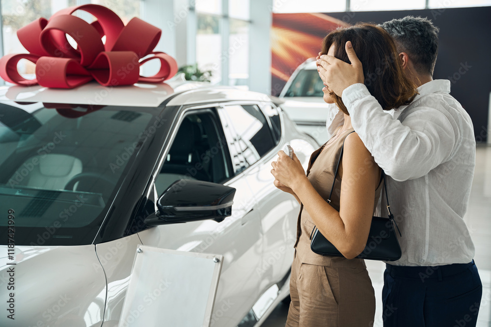 Man covering eyes of woman near new cars in modern auto salon