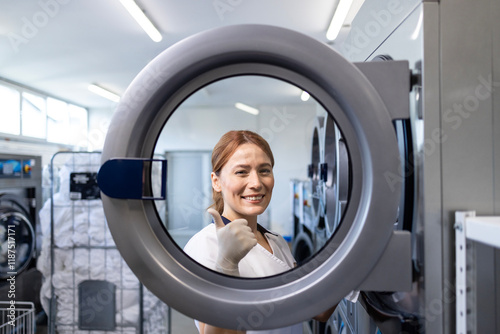 Female laundry worker standing by industrial washing machine and holding thumbs up in dry cleaning service. photo