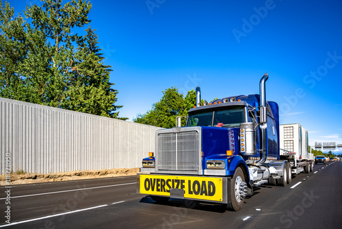 Bright blue big rig semi truck with oversize load sigh on the front bumper transporting oversized cargo on step deck semi trailer driving on the straight highway road at sunny day