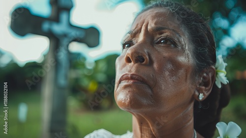 An elderly Hispanic woman gazes solemnly upward, her expression reflective and thoughtful, set against a blurred cross in the background. photo