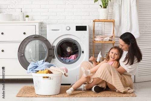 Young woman and her little daughter sitting on floor with laundry near washing machine at home photo