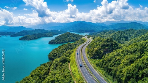 Aerial view of highway curving by lake with mountains, under cloudy blue sky