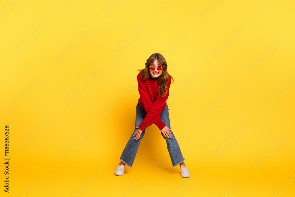 Young woman wearing fashionable red sweater and stylish sunglasses posing on vibrant yellow background, exuding charm and joy Young woman wearing fashionable red sweater and stylish sunglasses posing on vibrant yellow background, exuding charm and joy
