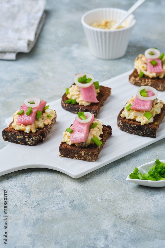 Appetizer, mini sandwiches on rye bread with egg salad, pickled herring and green onions on a white ceramic serving board on a gray concrete background. Sandwich recipes.