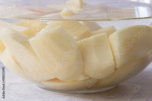 sliced potato soaking in a glass bowl of water. On a white marble background photo