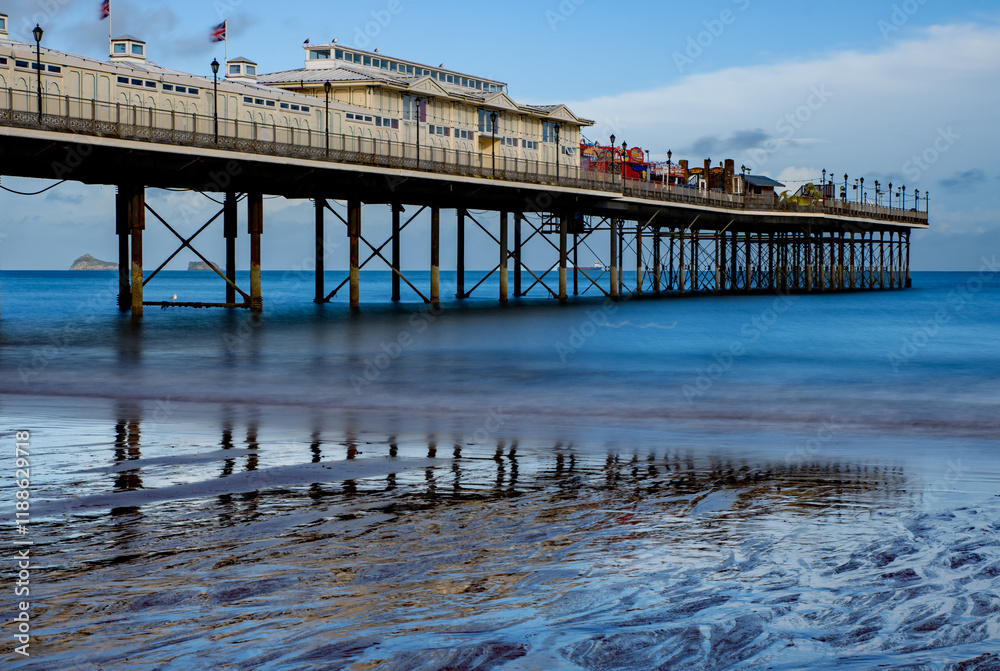 Fototapeta premium Seaside pier with reflections in the sand, long exposure with smooth sea, blue sky and sunshine. Dramatic, Sandy beach holiday resort. 
