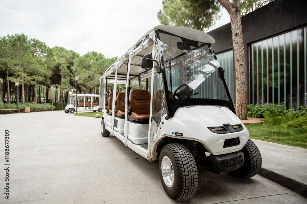 Obraz premium Close-up of a white electric cart with brown seats parked outdoors near trees and modern architecture, in a resort environment..
