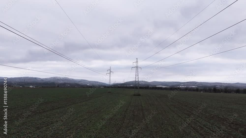 Transmission towers in a field with a moody, overcast sky. Drone footage glides along the power lines, capturing the industrial scene from above. Transmission towers in a field with a moody, overcast sky. Drone footage glides along the power lines, capturing the industrial scene from above.