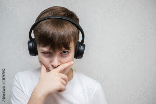 a ten year old boy looks at the camera and covers his mouth with his hand, thinking. in wireless headphones photo