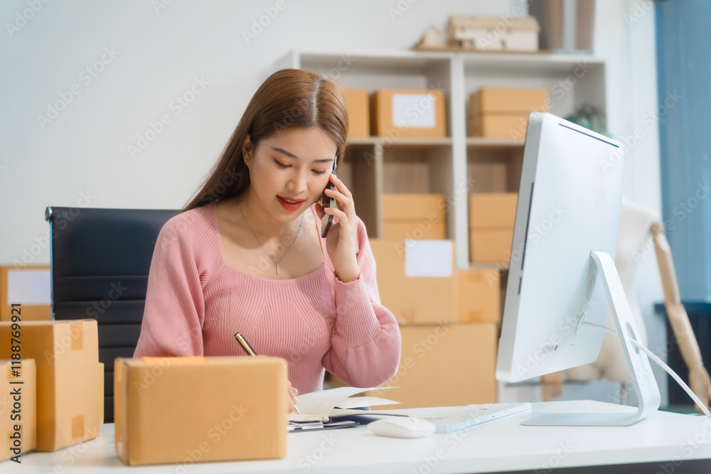 An Asian young woman sits in a room, working on a computer and making calls or video calls, managing her small business and online sales, surrounded by cardboard boxes ready for shipping. An Asian young woman sits in a room, working on a computer and making calls or video calls, managing her small business and online sales, surrounded by cardboard boxes ready for shipping.