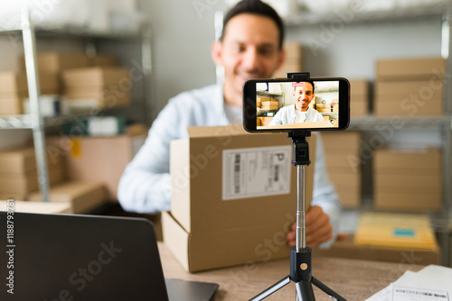 Smiling business owner with a package, creating a promotional video for his online store using a smartphone