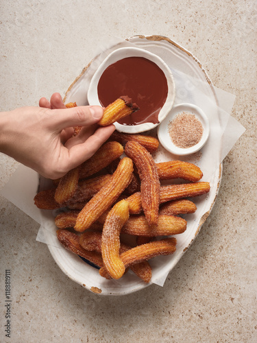 A hand reaches for a crispy churro, ready to dip into rich chocolate sauce. Cinnamon sugar sits nearby, enticing sweet lovers in a cozy setting photo